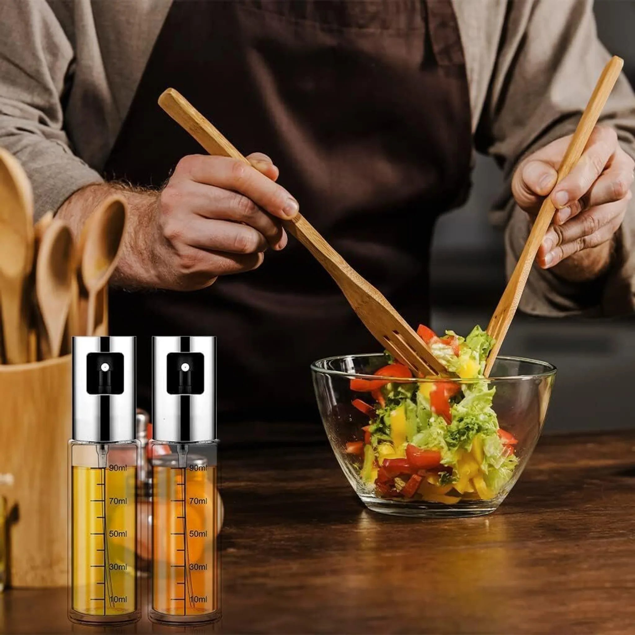 Eine Person wirft Salat in einer Glasschüssel mit Holzutensilien. Ölsprühflaschen sind im Vordergrund.