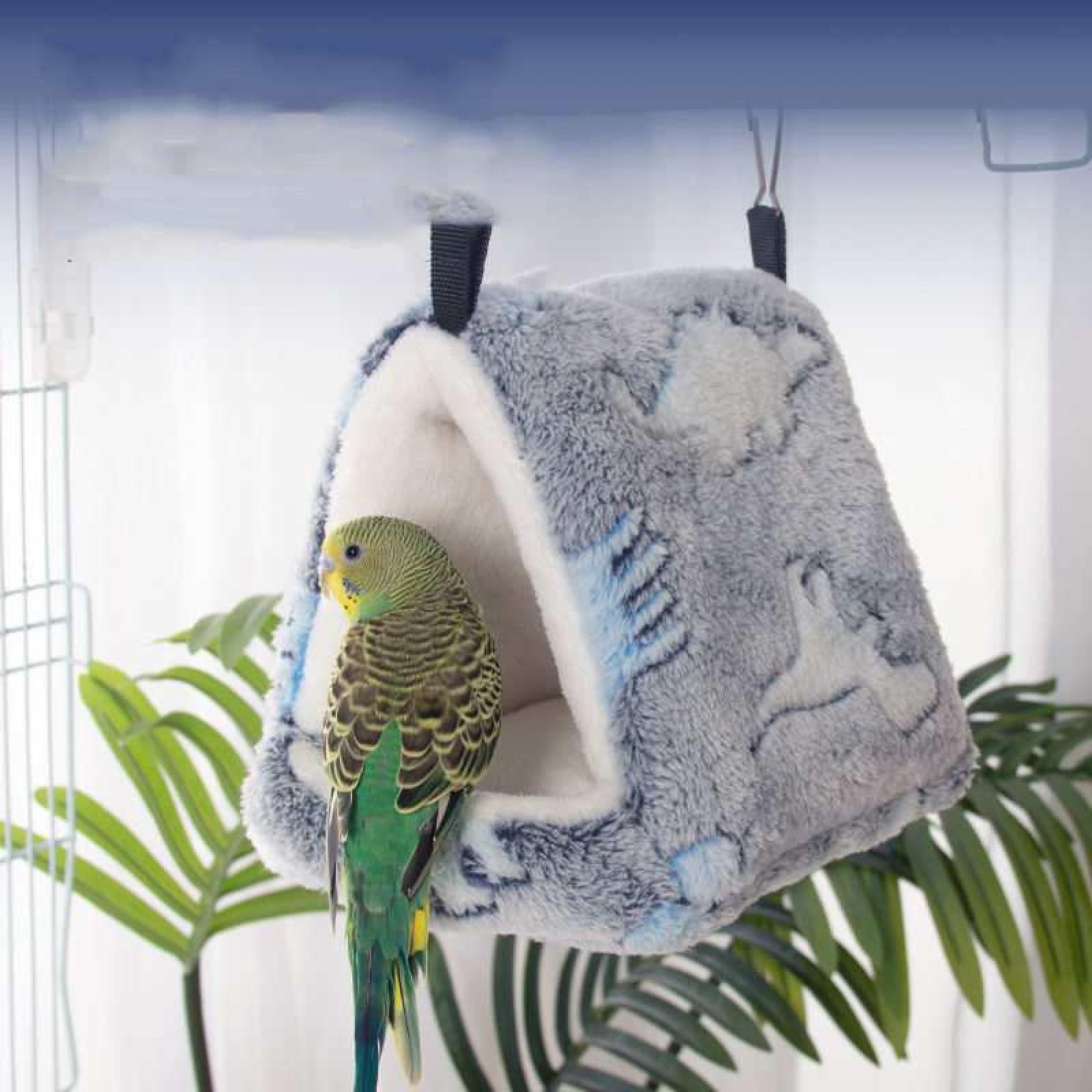 Ein grüner Wellensittich sitzt in der Nähe eines flauschigen, grauen Vogelhauses. Im Hintergrund befinden sich grüne Blätter. Das Vogelhaus hängt.