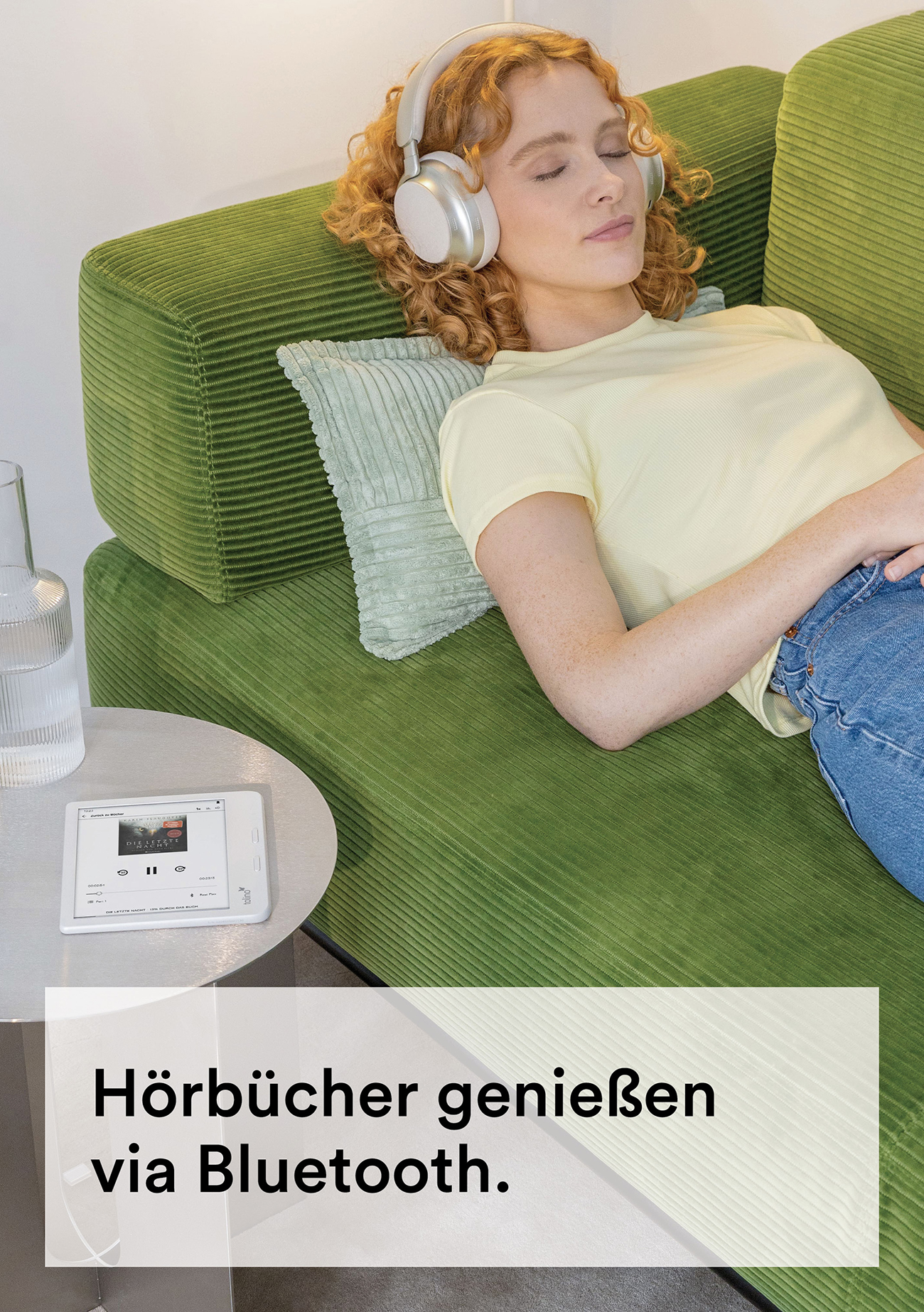 Una donna con le cuffie si rilassa su un divano verde, ascoltando un libro su un tablet. Un bicchiere e un cuscino sono vicini.