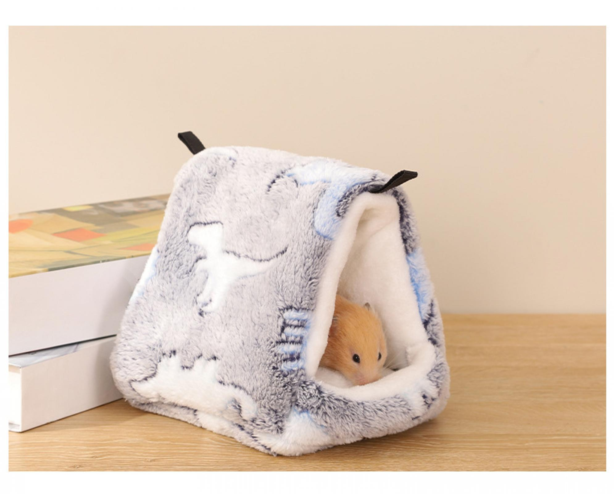 Ein Hamster in einem weichen grau-weißen Bett. Das Bett steht auf einem Holztisch. Der Hintergrund ist eine Wand und Bücher.