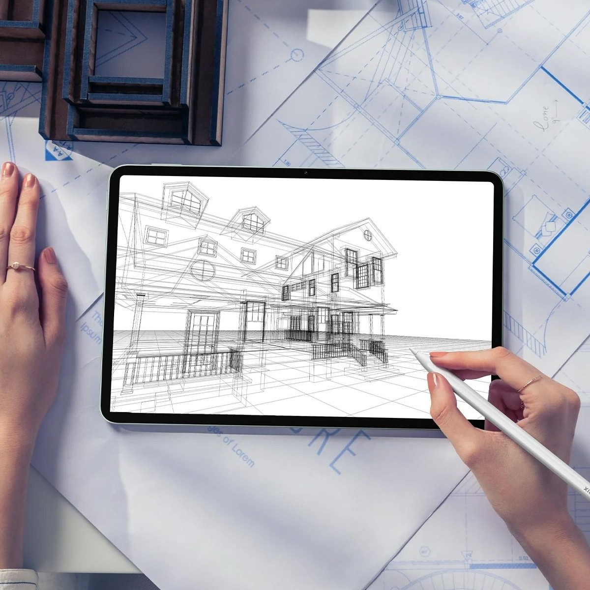 Ein Tablet zeigt einen Hausentwurf. Hände mit Ringen halten Tablet und Stift. Blaupausen und ein Modellhaus im Hintergrund.