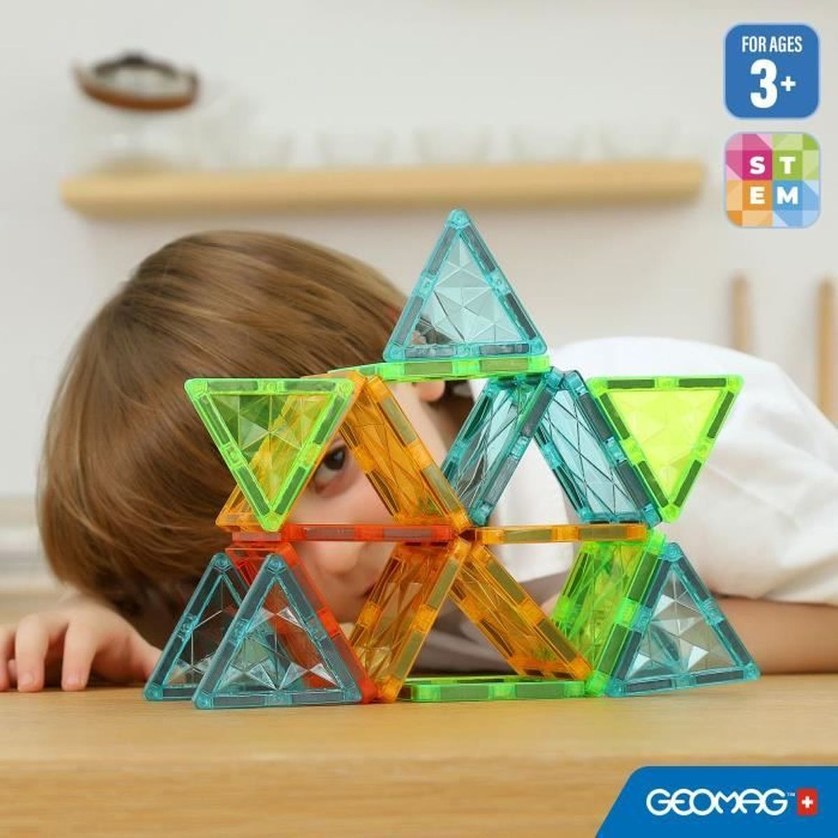 Ein Kind betrachtet ein farbenfrohes magnetisches Konstruktionsspielzeug aus Dreiecken. Der Hintergrund ist verschwommen und zeigt einen Tisch und Regale.