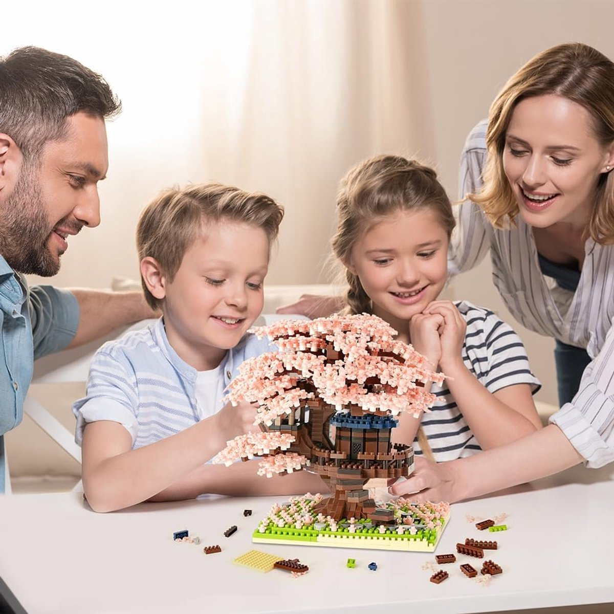 Eine Familie baut einen Spielzeugbaum mit rosa Blüten. Ein Junge und ein Mädchen bauen ihn mit ihren Eltern an einem weißen Tisch.