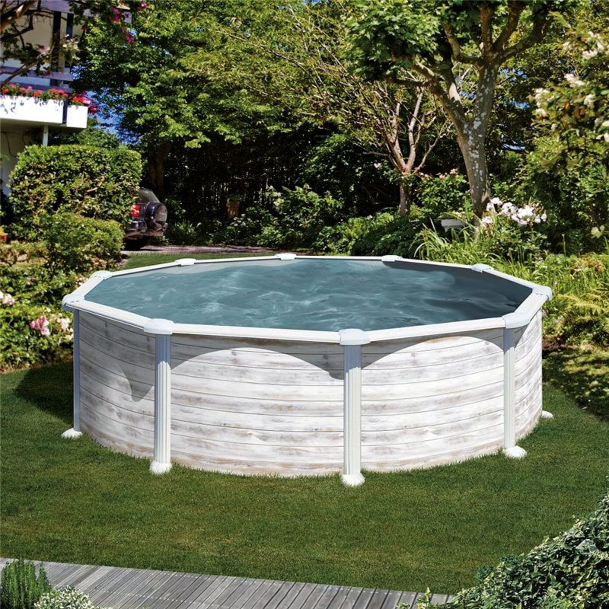 Una piscina elevada se encuentra sobre hierba. Árboles y arbustos la rodean. La piscina tiene lados blancos y agua azul.