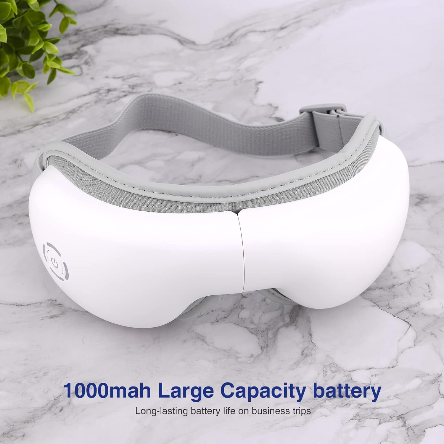 Ein weißes Augenmassagegerät mit grauem Riemen. Es liegt auf einer Marmoroberfläche. Text: 1000mAh Große Kapazität Batterie.