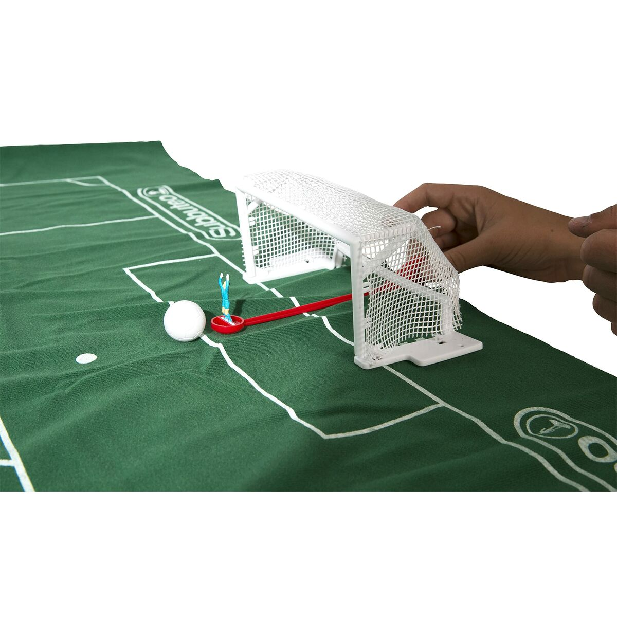 Configuración de un juego de fútbol en miniatura. Una portería blanca, un balón, una figura de jugador y un palo rojo sobre un campo verde. Una mano se acerca.