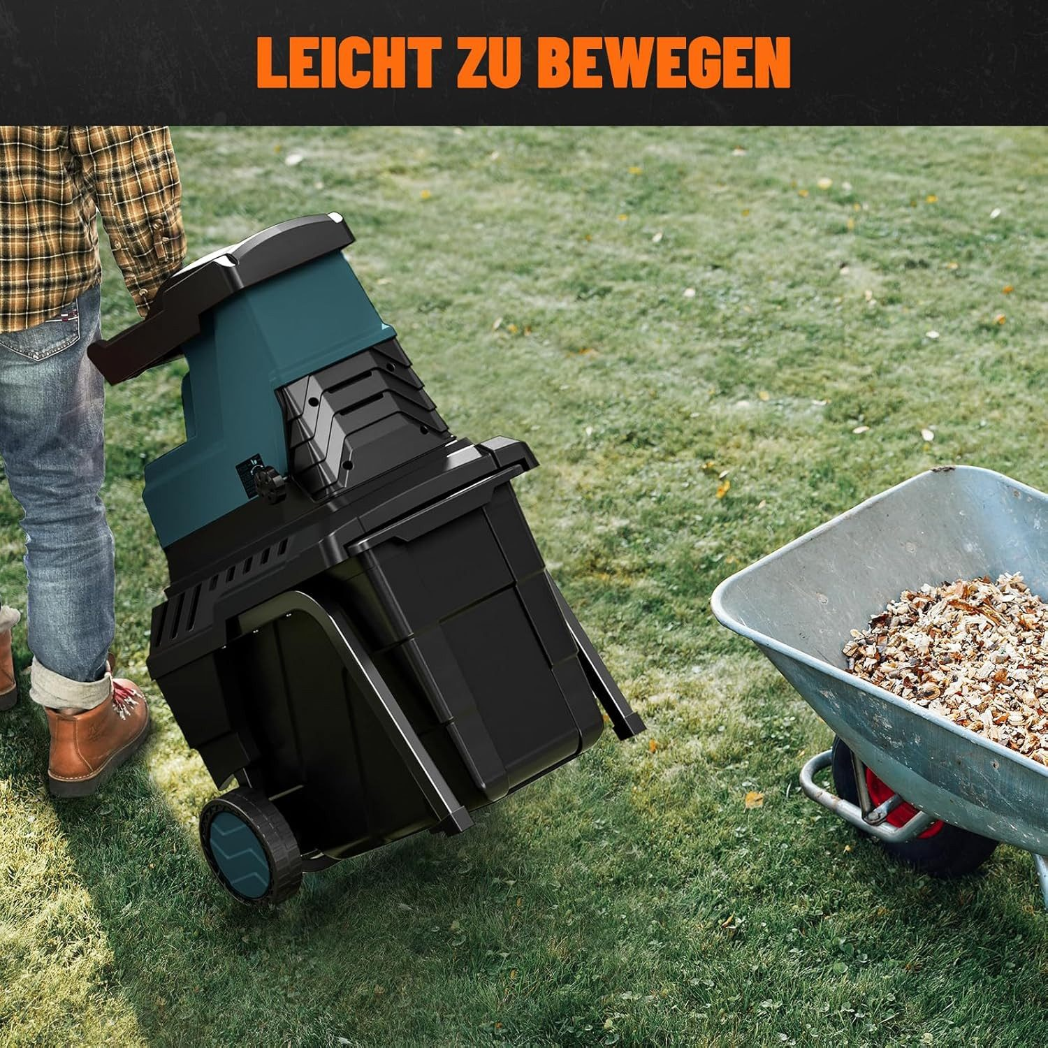 Eine Person bewegt einen schwarzen und tealfarbenen Holzhäcksler auf Gras. Eine Schubkarre mit Holzhackschnitzeln steht daneben.