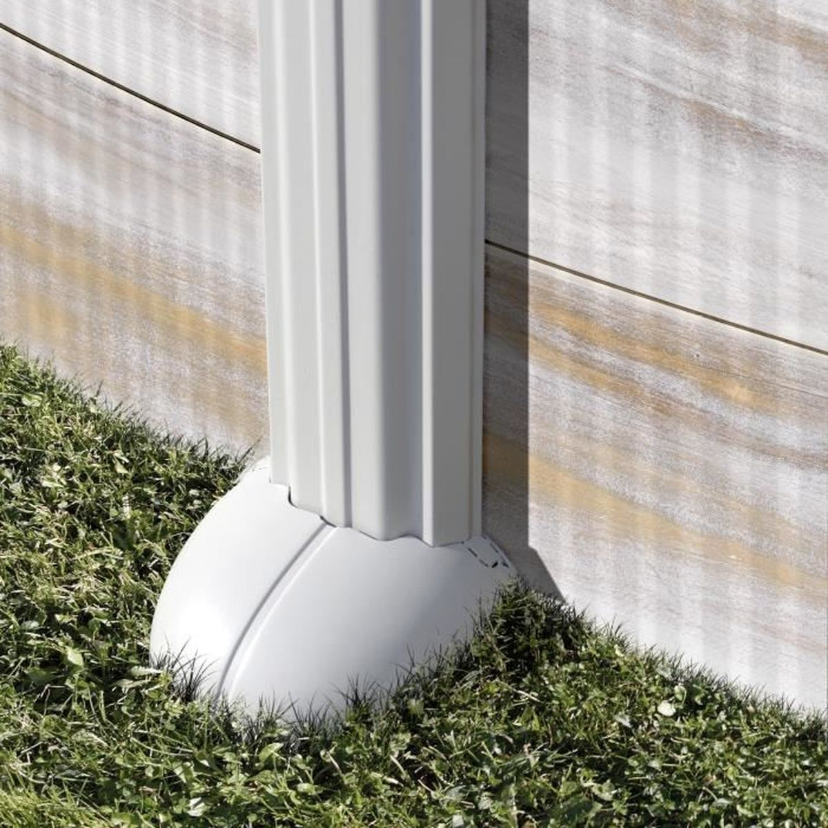 Una tubería blanca de canaleta con base redondeada se asienta sobre hierba verde. Una pared de madera en el fondo.