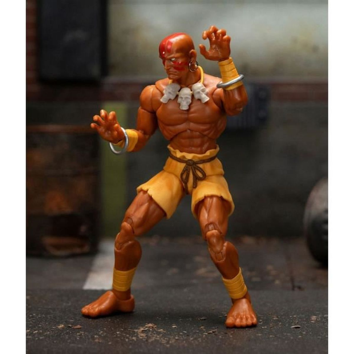 Actionfigur eines glatzköpfigen Mannes mit roten Markierungen, der gelbe Shorts und Armbänder sowie eine Totenkette trägt und eine Pose einnimmt.
