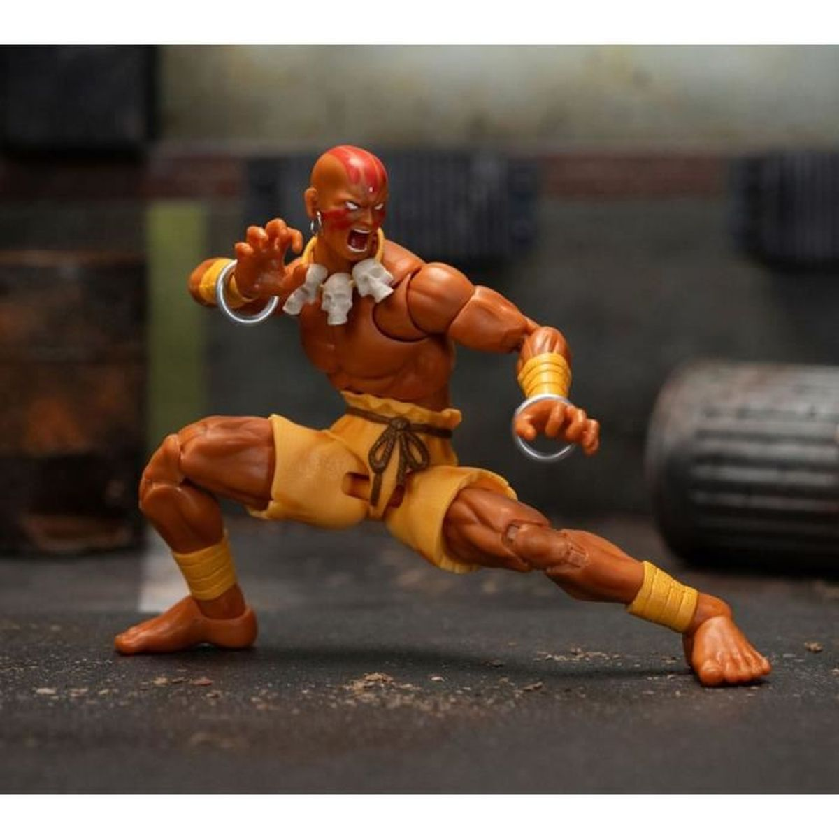 Actionfigur eines glatzköpfigen Mannes in Kampfpose, mit gelben Shorts und Armbändern. Er hat rote Gesichtsbemalung und weiße Halsketten.
