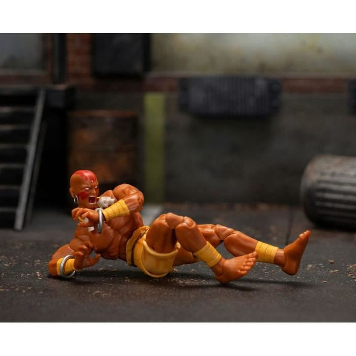 Eine kahle Actionfigur in Kampfpose, mit gelben Arm- und Beinwickeln. Die Figur liegt am Boden, im Hintergrund eine Backsteinmauer.