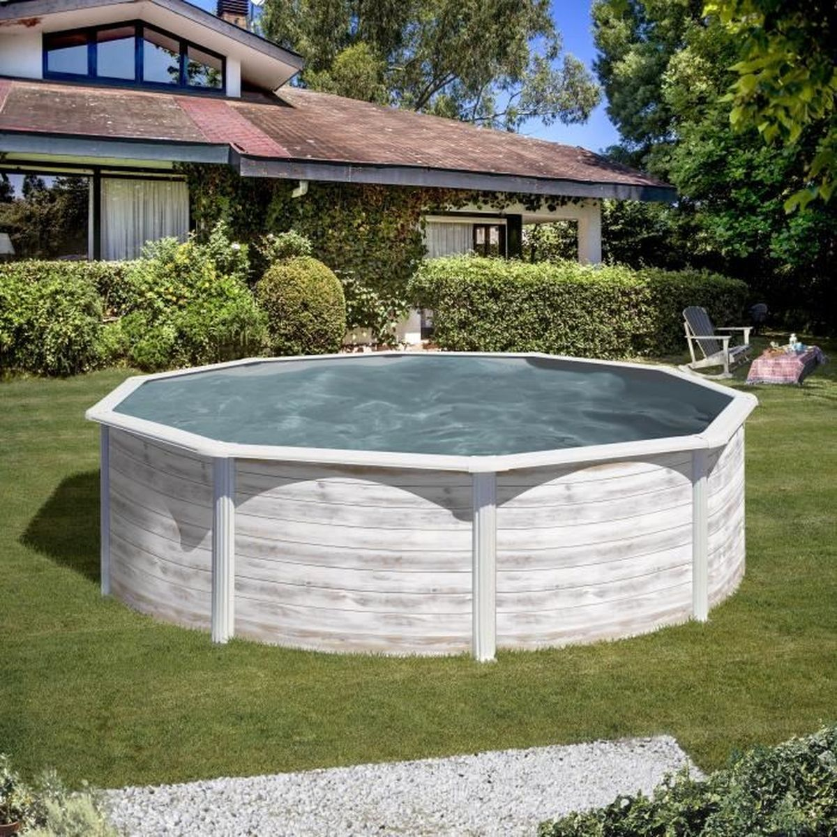 Una piscina redonda de madera llena de agua está sobre hierba verde, con una casa y árboles al fondo bajo un cielo azul.