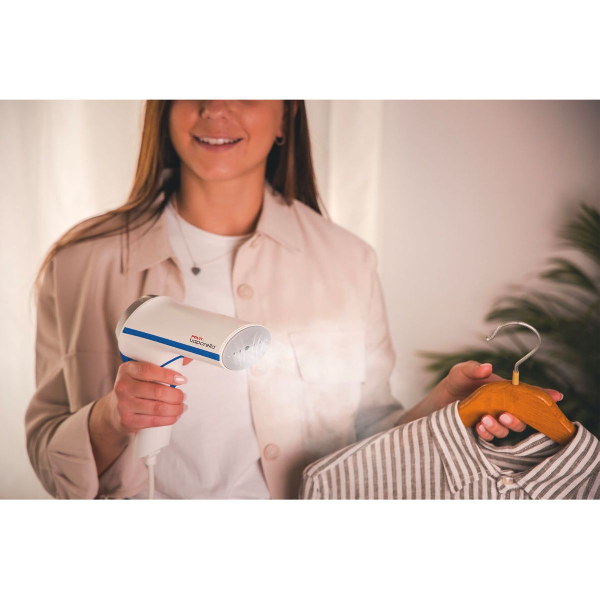 Una donna stira una camicia con un vaporizzatore Polti Vaporella. La camicia è appesa a una gruccia di legno. Il vapore è visibile. Lo sfondo è sfocato.