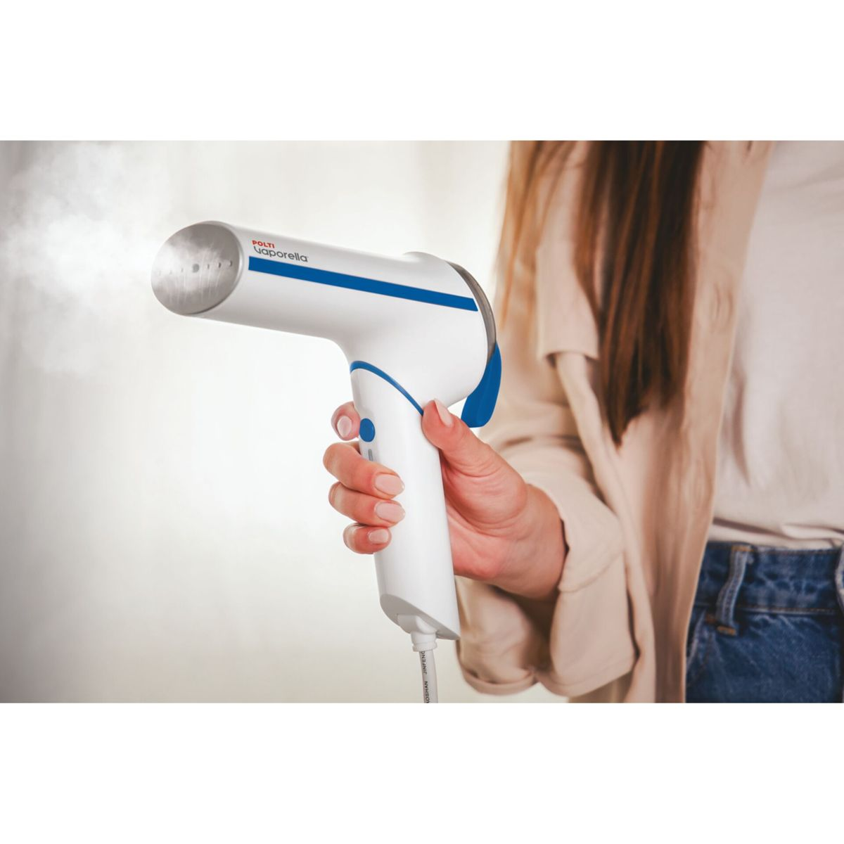 Una donna tiene in mano un vaporizzatore per indumenti bianco e blu, il vapore esce dall'ugello. Indossa una maglia beige e jeans.