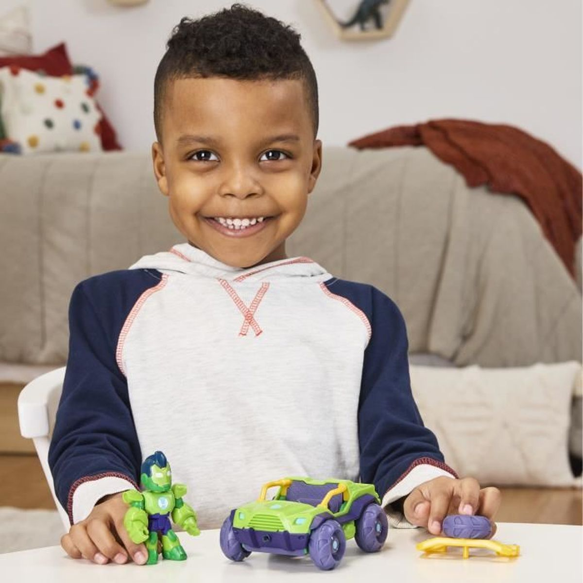 Un niño sonriente sostiene juguetes: una figura verde, un coche verde y morado y un juguete amarillo. El fondo muestra una cama y un dinosaurio.