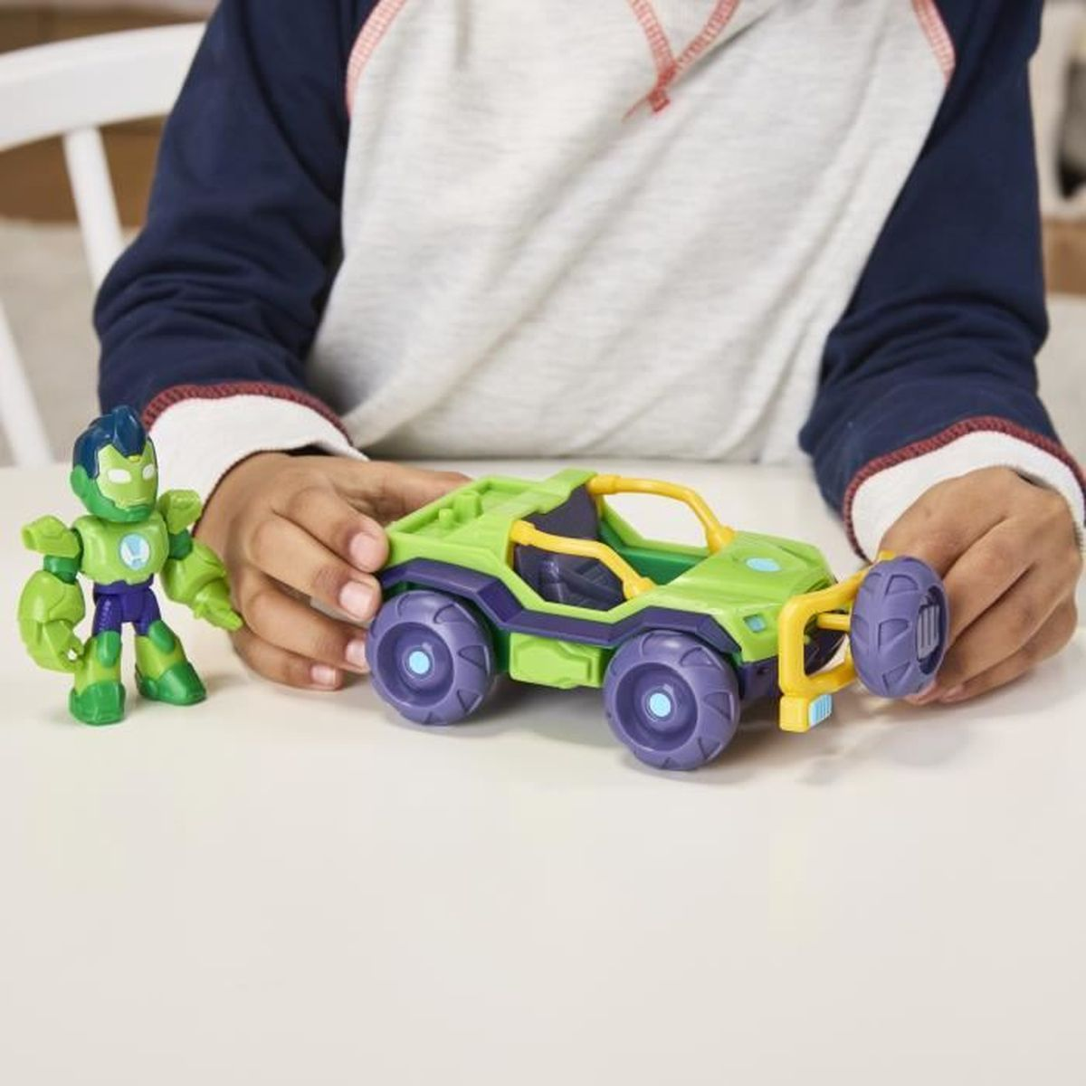 Las manos de un niño juegan con un coche de juguete verde y una figura de superhéroe verde, sobre una superficie blanca.