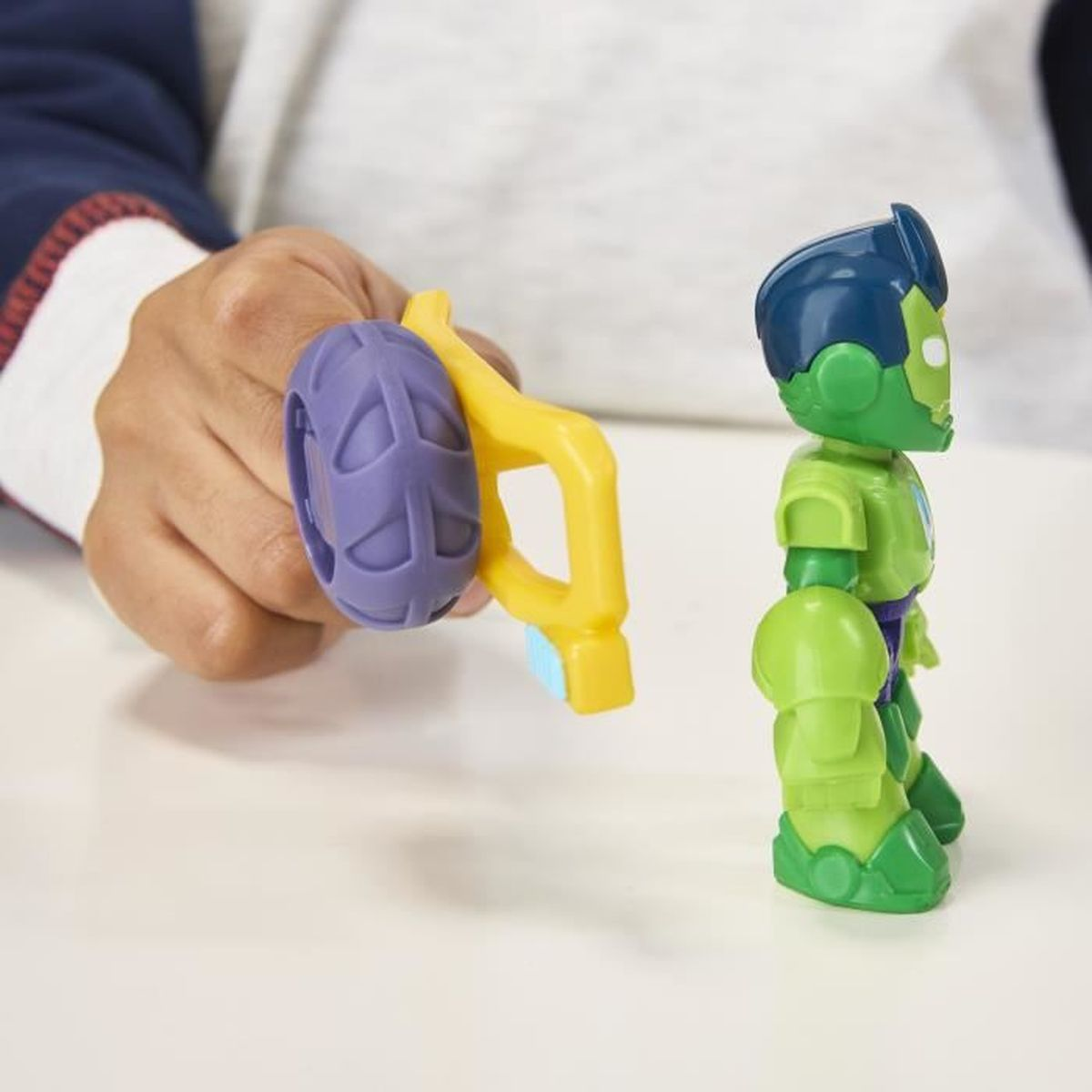 Una mano sostiene un juguete amarillo con una rueda morada. Un robot de juguete verde con pelo azul está de pie sobre una superficie blanca.