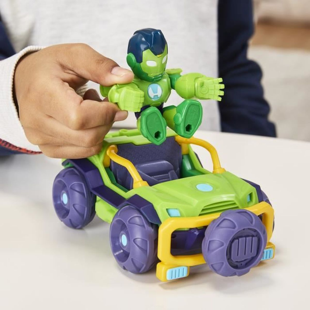 La mano de un niño sostiene una figura de juguete verde de Iron Man sentada en un coche de juguete verde y morado con detalles amarillos. El fondo está borroso.