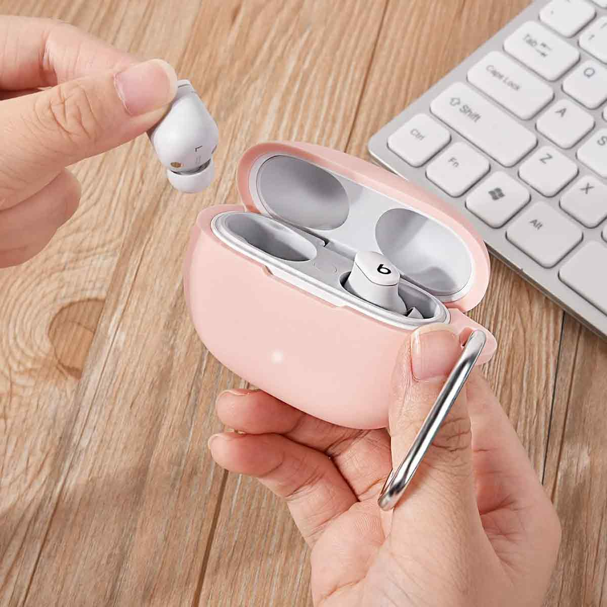 Eine Hand hält einen weißen Ohrhörer, eine andere Hand hält ein rosafarbenes Etui mit einem Ohrhörer darin, in der Nähe einer weißen Tastatur.