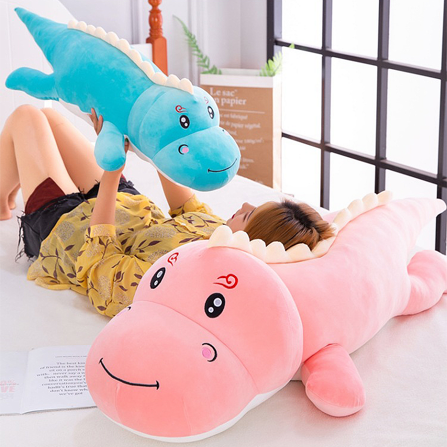 Una persona yace en una cama, sosteniendo un dinosaurio de peluche azul. Un dinosaurio de peluche rosa está al frente. El fondo incluye una ventana y una bolsa de papel.