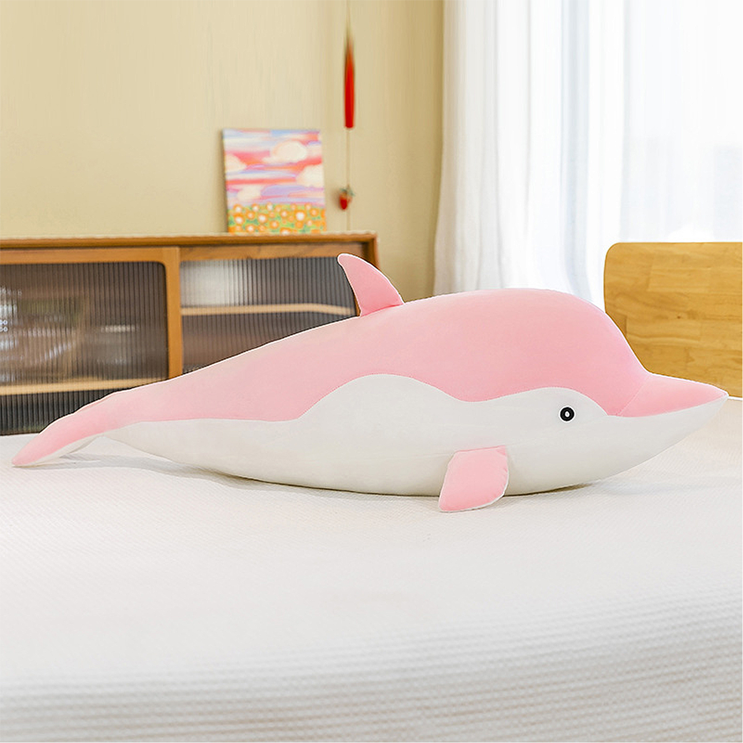 Un peluche a forma di delfino rosa e bianco su un letto bianco. Un mobile in legno e un dipinto sullo sfondo.