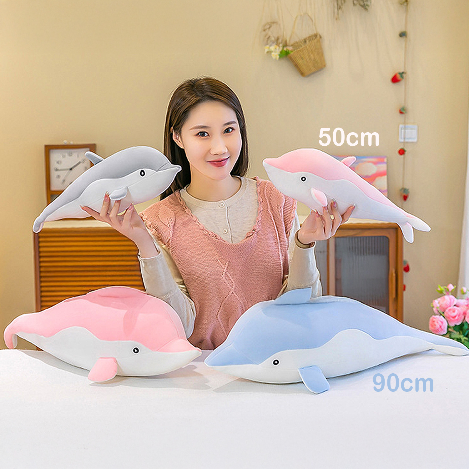 Donna che tiene tre peluche di delfino: grigio, rosa e rosa/blu. Un grande delfino blu è sul tavolo. Un orologio e fiori sullo sfondo.