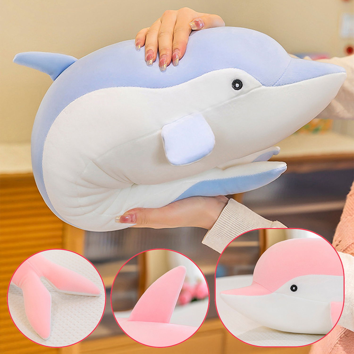 Un morbido peluche a forma di delfino blu e bianco è tenuto da una persona. Dettagli rosa sono visibili sulle pinne e sugli inserti. Vengono mostrate viste aggiuntive.