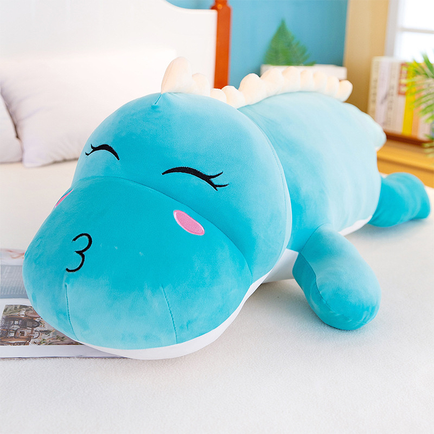 Un dinosaurio de peluche azul con los ojos cerrados y el número 3 en la cara, descansando sobre una superficie blanca.