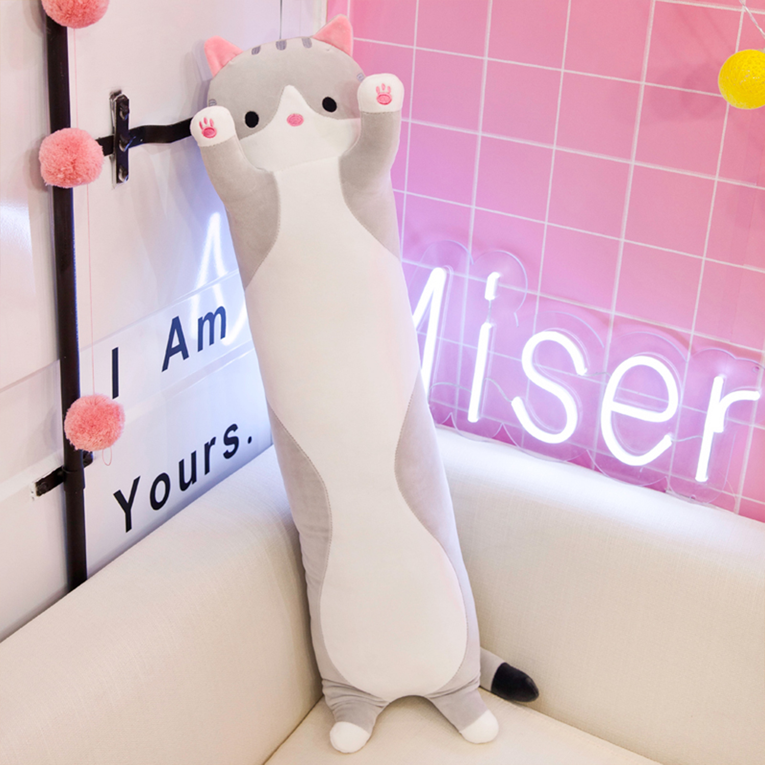 Un lungo peluche a forma di gatto è appoggiato a un muro. Il muro è rosa e ha un'insegna al neon che dice 'Miser'. Il gatto è su un divano bianco.