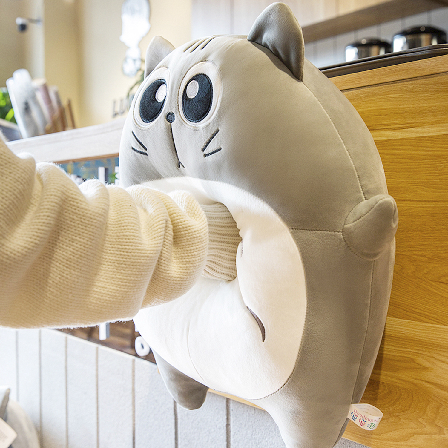Una mano con un maglione color crema interagisce con un peluche grigio a forma di gatto. Il peluche ha grandi occhi e si trova su una superficie di legno.