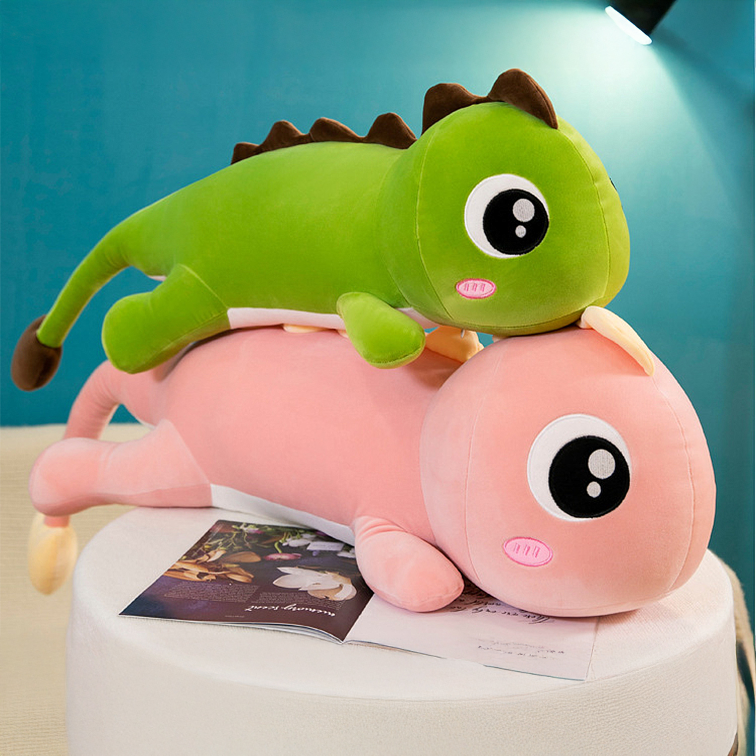 Due dinosauri di peluche, verde sopra rosa, su una superficie bianca con una rivista. Sfondo blu.