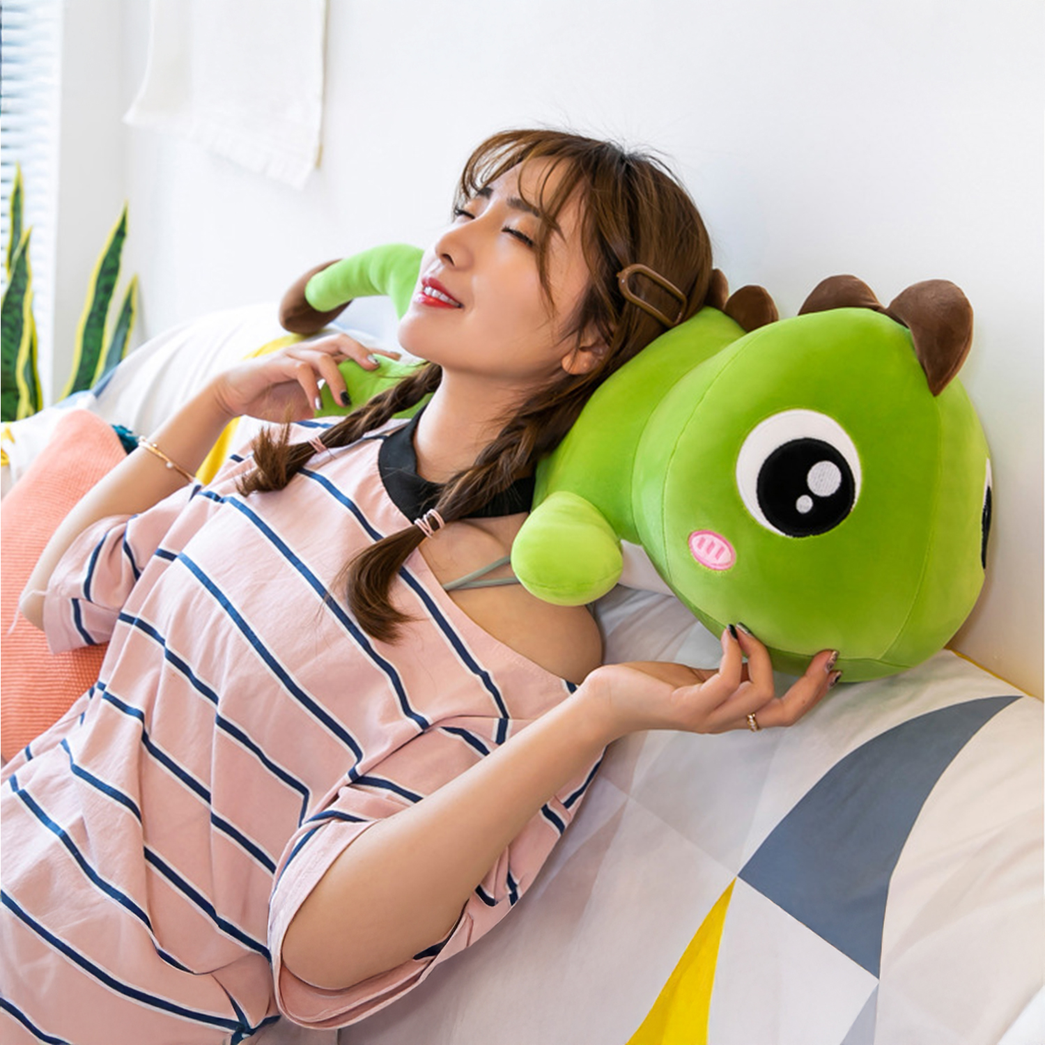 Una donna riposa su un letto, tenendo in mano un peluche di dinosauro verde. Indossa una camicia a righe. La scena è luminosa e al chiuso.