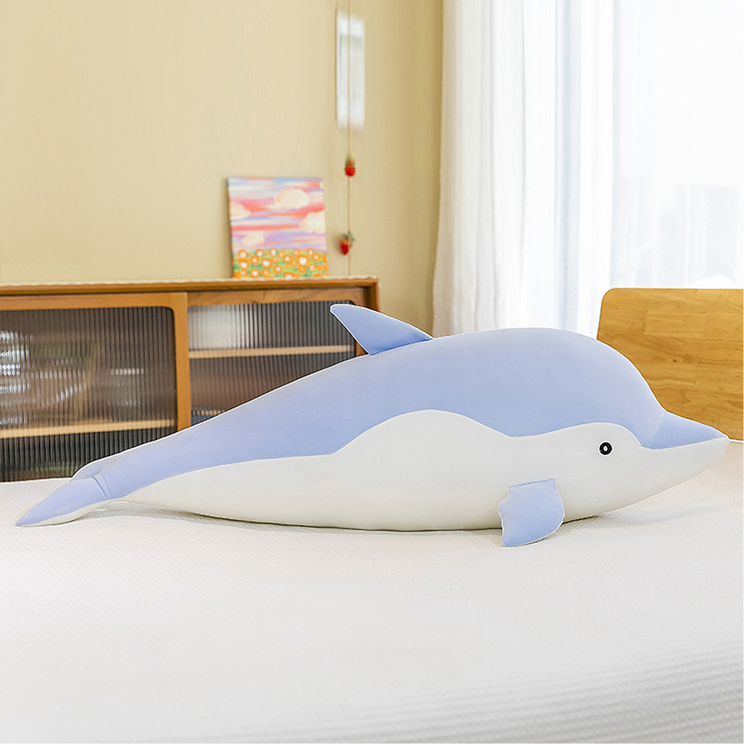 Un delfino di peluche blu e bianco giace su un letto bianco. Lo sfondo include un armadio in legno, opere d'arte e una finestra.