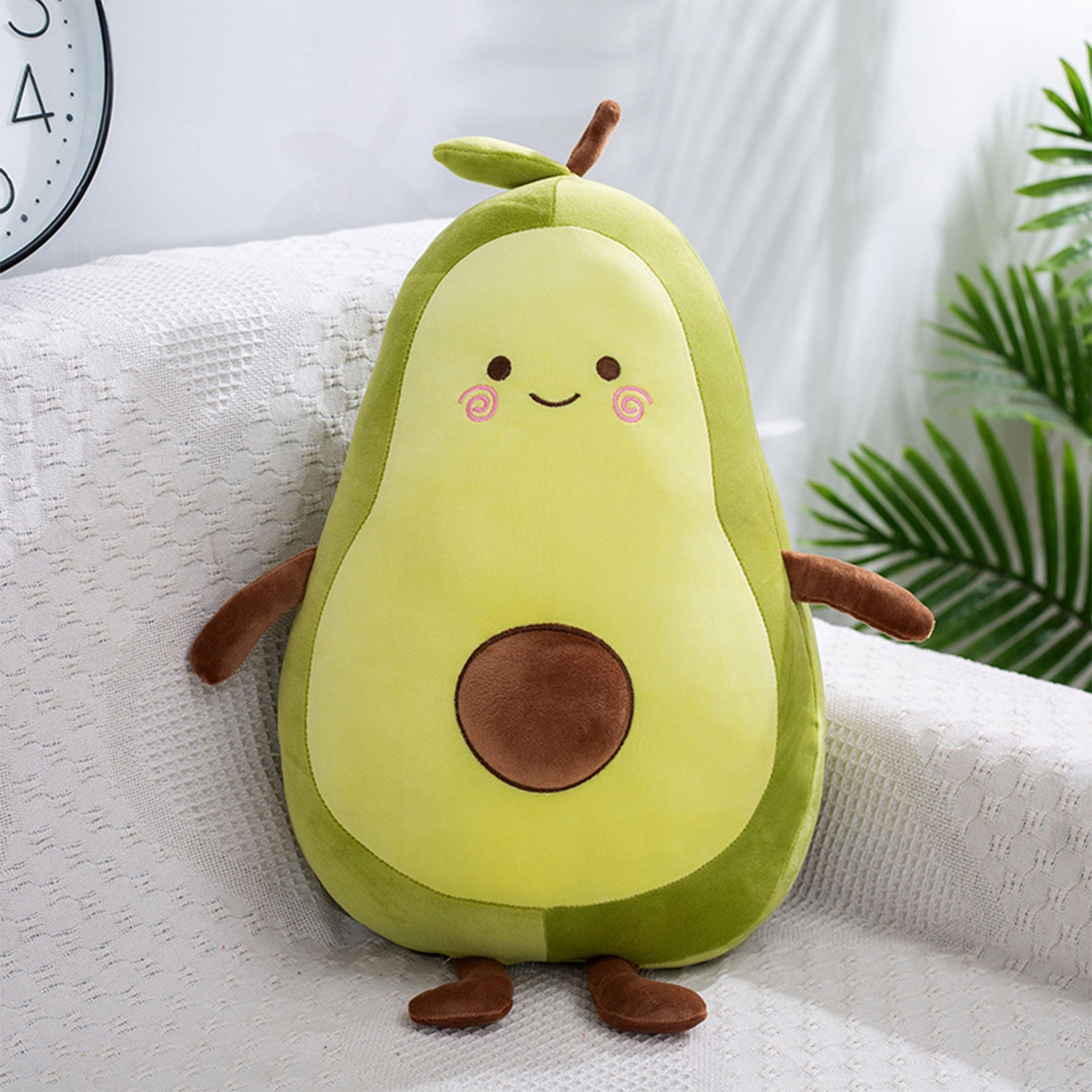 Un giocattolo di avocado di peluche con un viso sorridente, braccia e piedi. È seduto su un divano bianco a fantasia, con un orologio e una pianta.