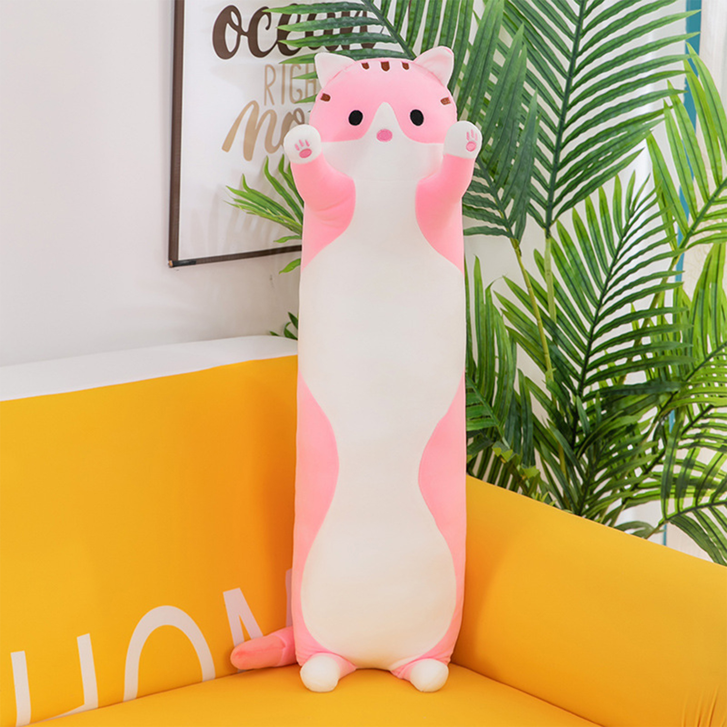 Un largo peluche de gato rosa en un sofá amarillo. Detrás hay un cuadro y plantas verdes. El gato tiene las patas levantadas.