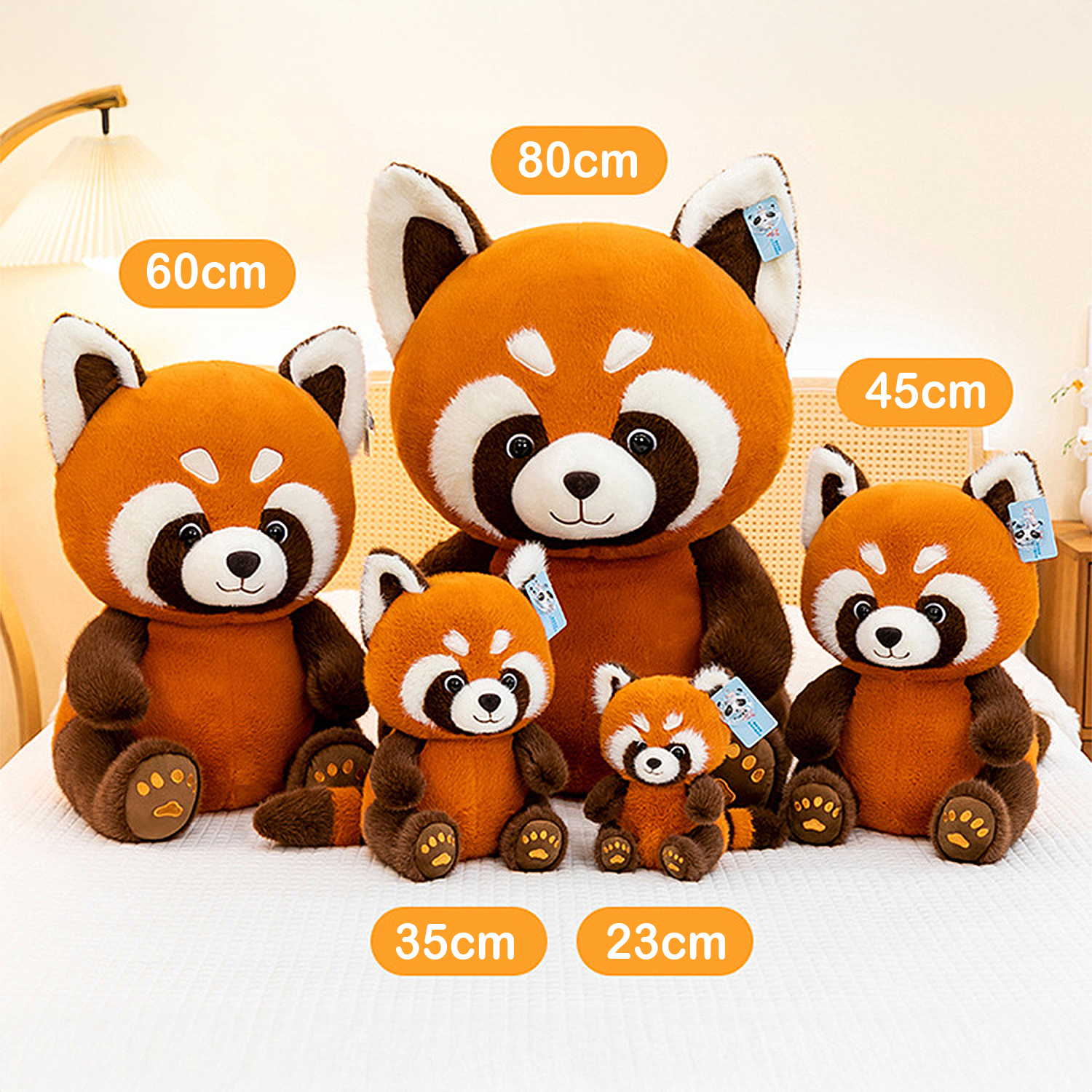 Cinque peluche di panda rosso di diverse dimensioni sono disposti su un letto bianco. Ogni giocattolo ha un'etichetta. Le dimensioni sono indicate.