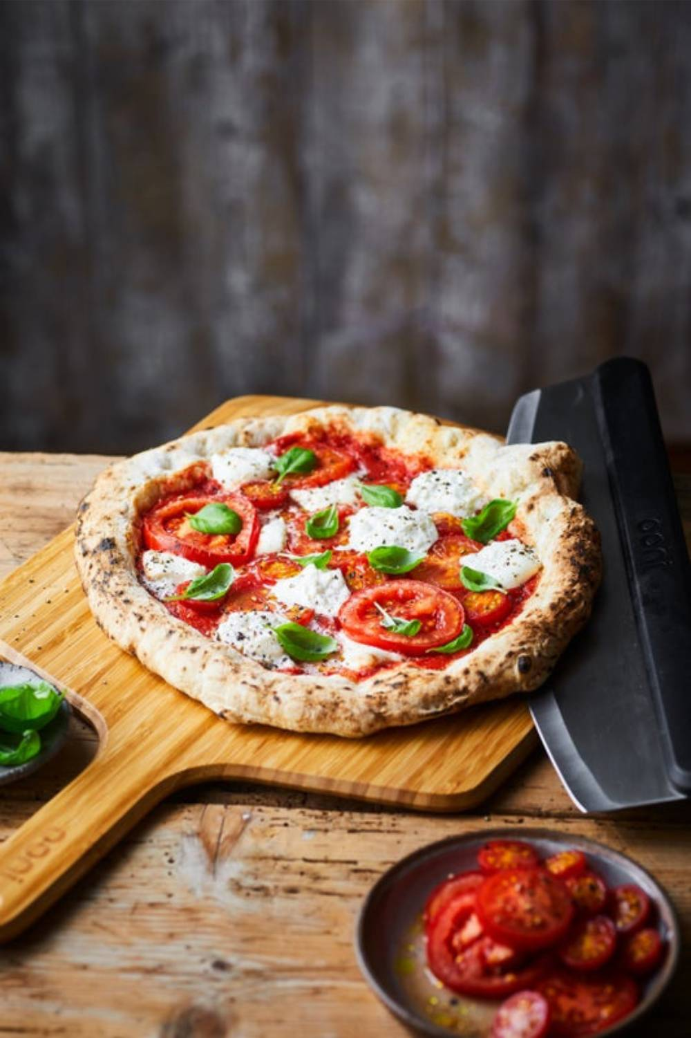 Eine Pizza mit Tomatenscheiben, Mozzarella und Basilikum auf einem Holzbrett, mit Pizzaschneider und Tomaten.