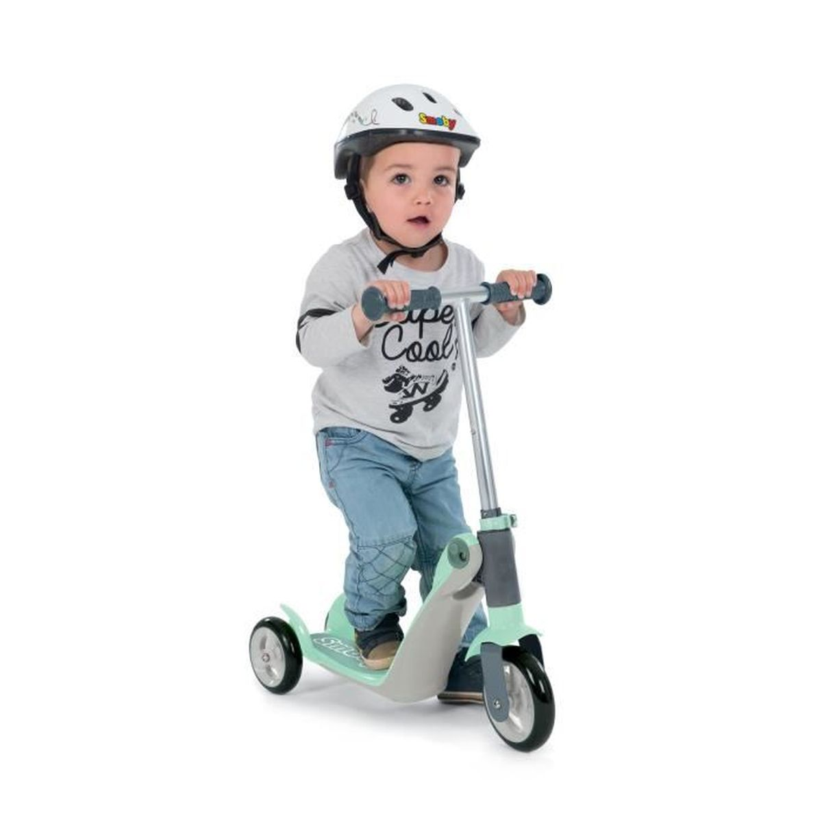 Ein Kleinkind mit Helm fährt einen grünen Roller. Das Kind trägt ein graues Hemd und Jeans. Weißer Hintergrund.