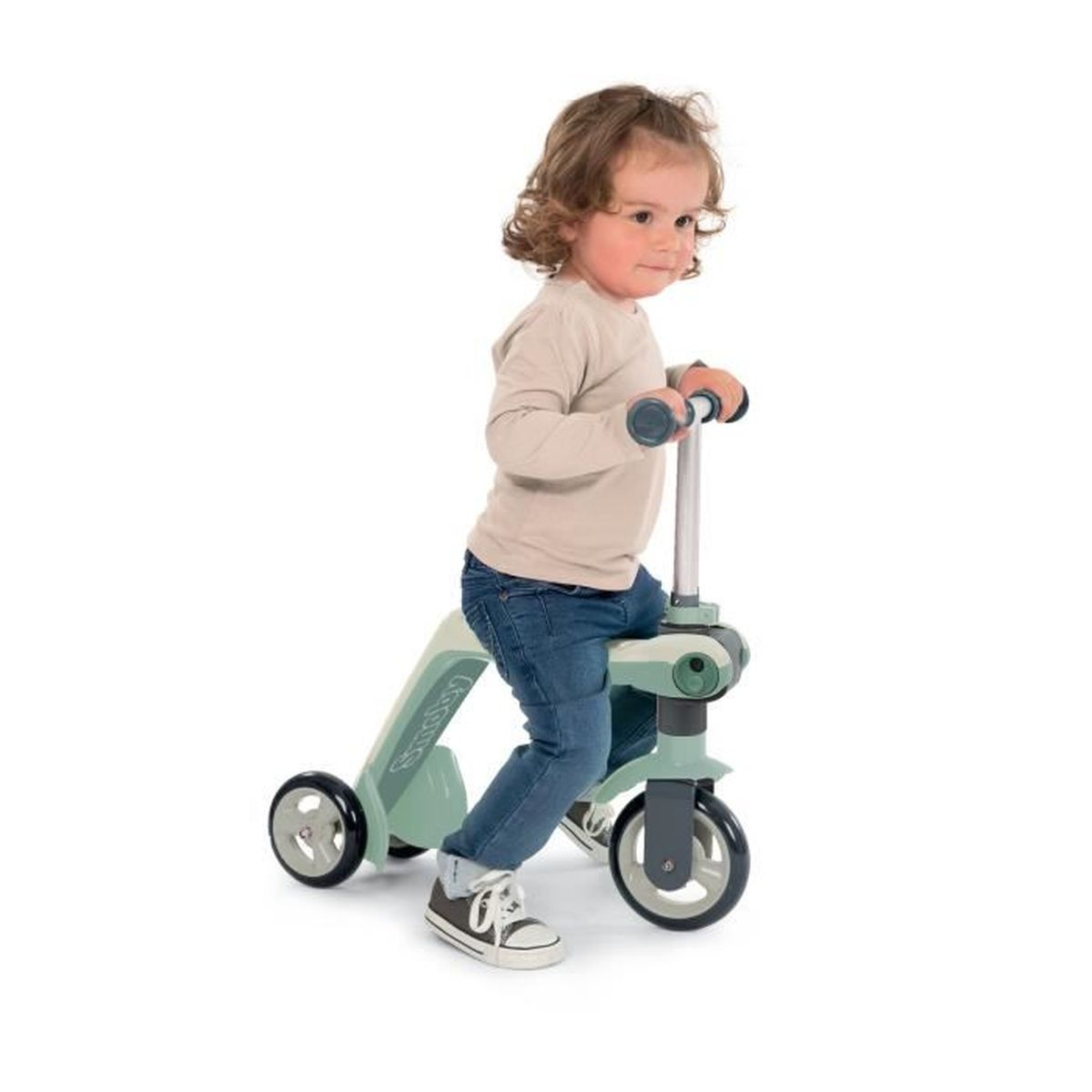 Ein Kleinkind fährt einen grünen Roller. Das Kind trägt ein beiges Hemd, Jeans und Turnschuhe. Der Roller hat drei Räder.