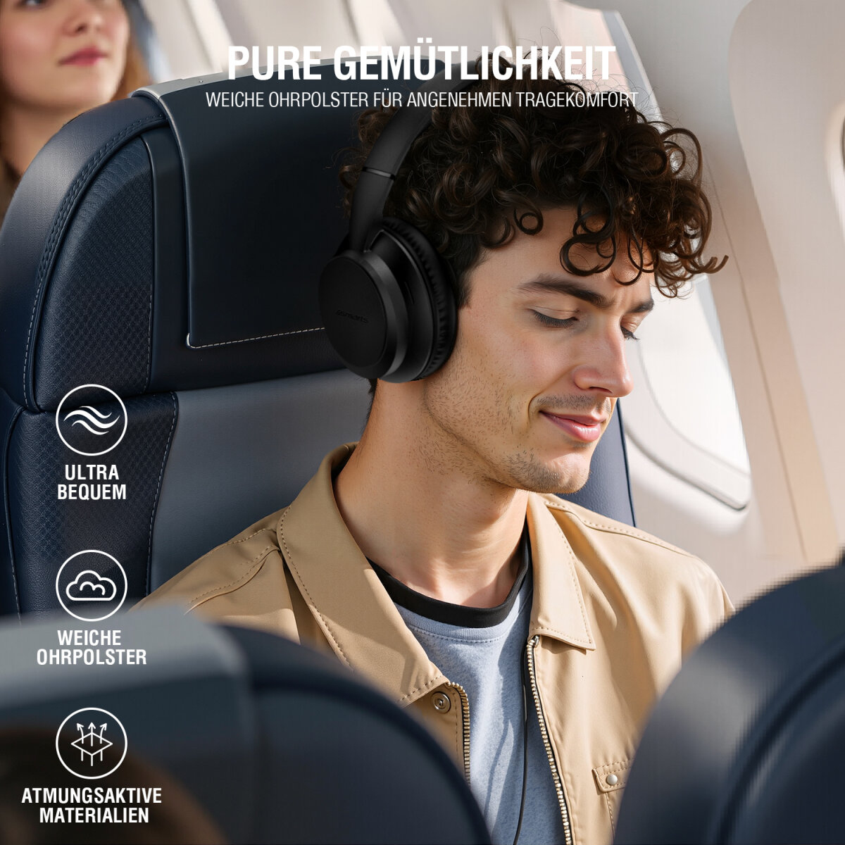 Un uomo con cuffie nere è in aereo, rilassato. Indossa una giacca beige. Un'altra persona è sullo sfondo.