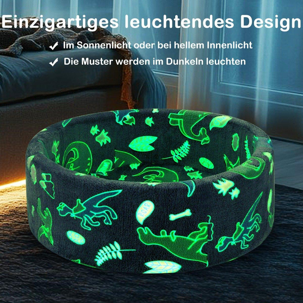 Una cama brillante para perros con patrones de dinosaurios. La cama es gris oscuro y los dinosaurios brillan en verde. El fondo incluye un sofá y una ventana.