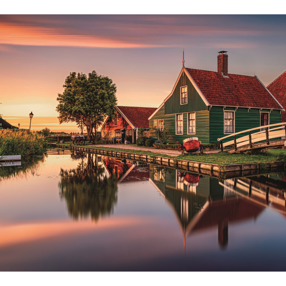 Zielone domy z czerwonymi dachami odbijają się w spokojnej wodzie. W pobliżu znajduje się mały most i drzewo. Niebo jest kolorowe o zachodzie słońca.