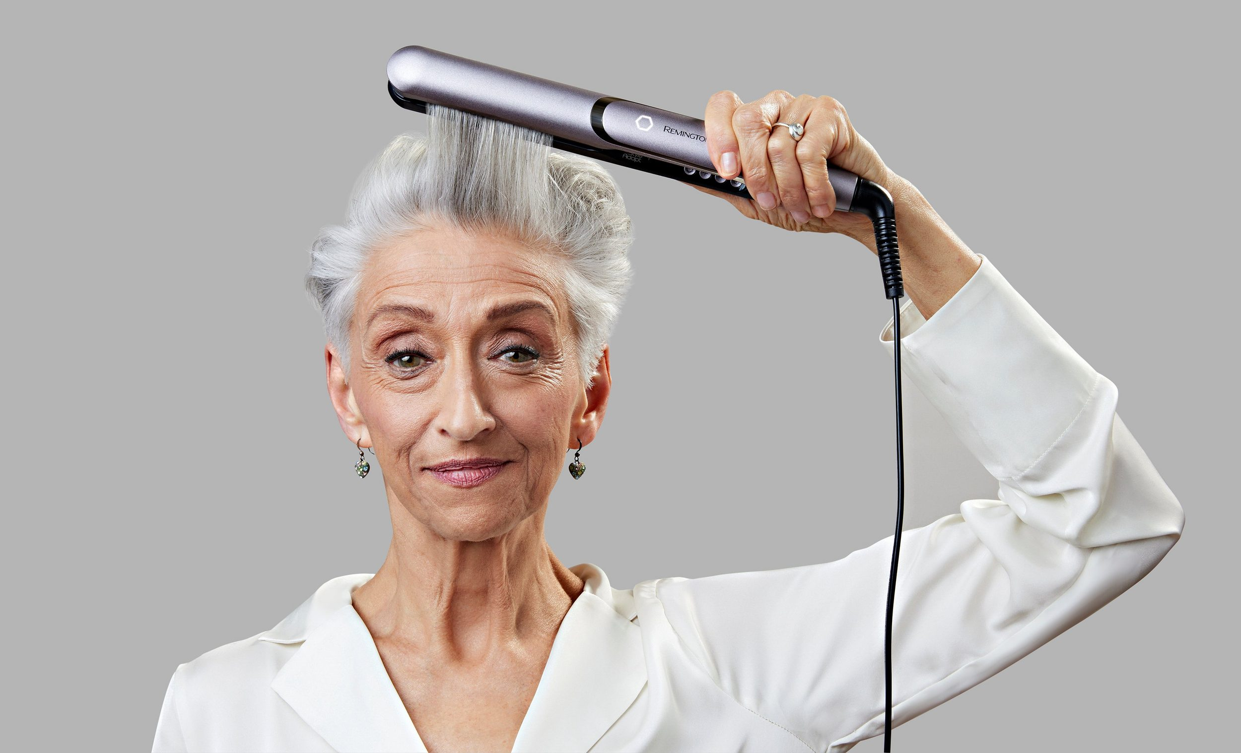 Una donna anziana usa una piastra per capelli. Ha i capelli bianchi, indossa orecchini e un top bianco. Sfondo grigio.