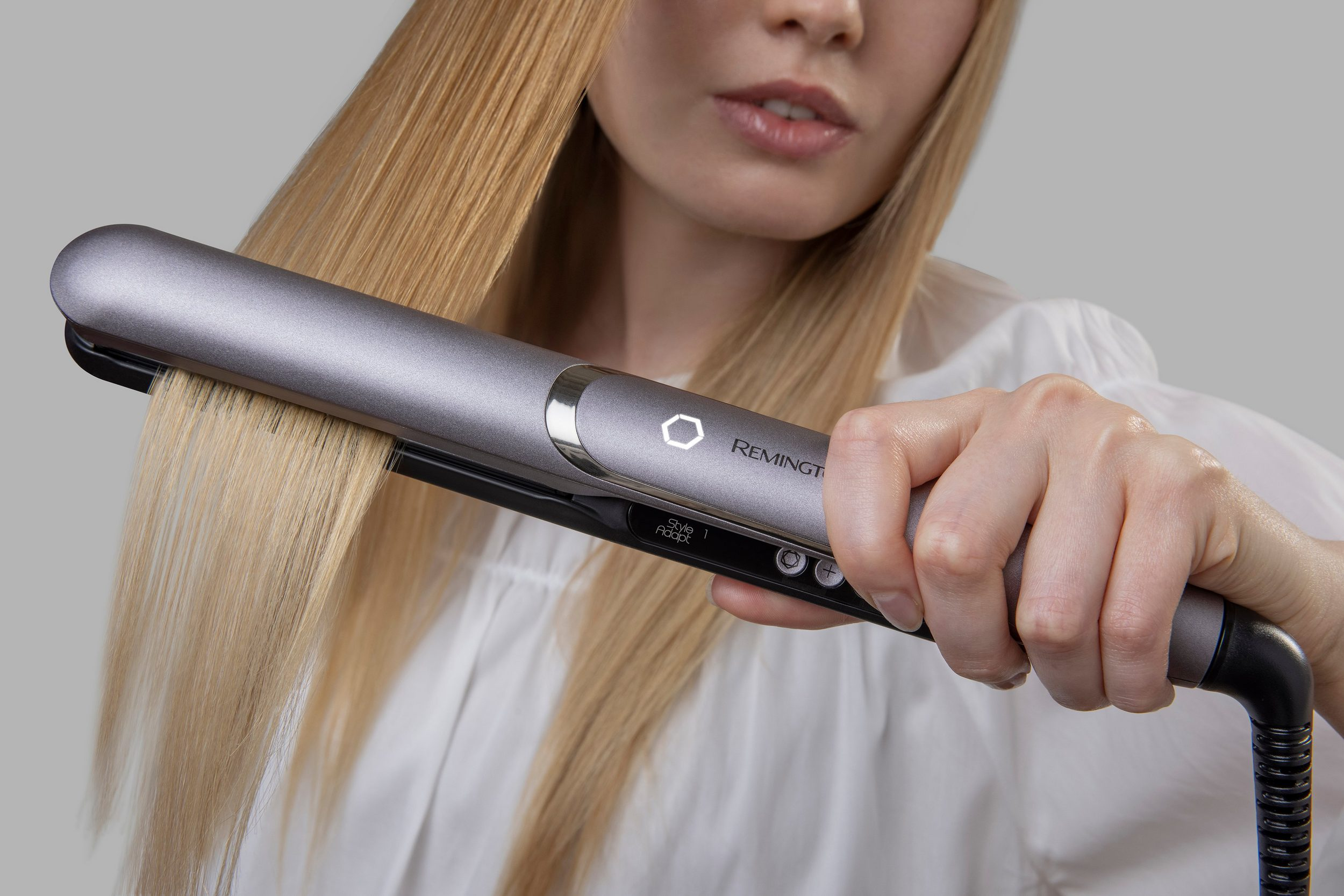 Una donna usa una piastra per capelli biondi. Il dispositivo è grigio e Remington. Indossa una camicia bianca.