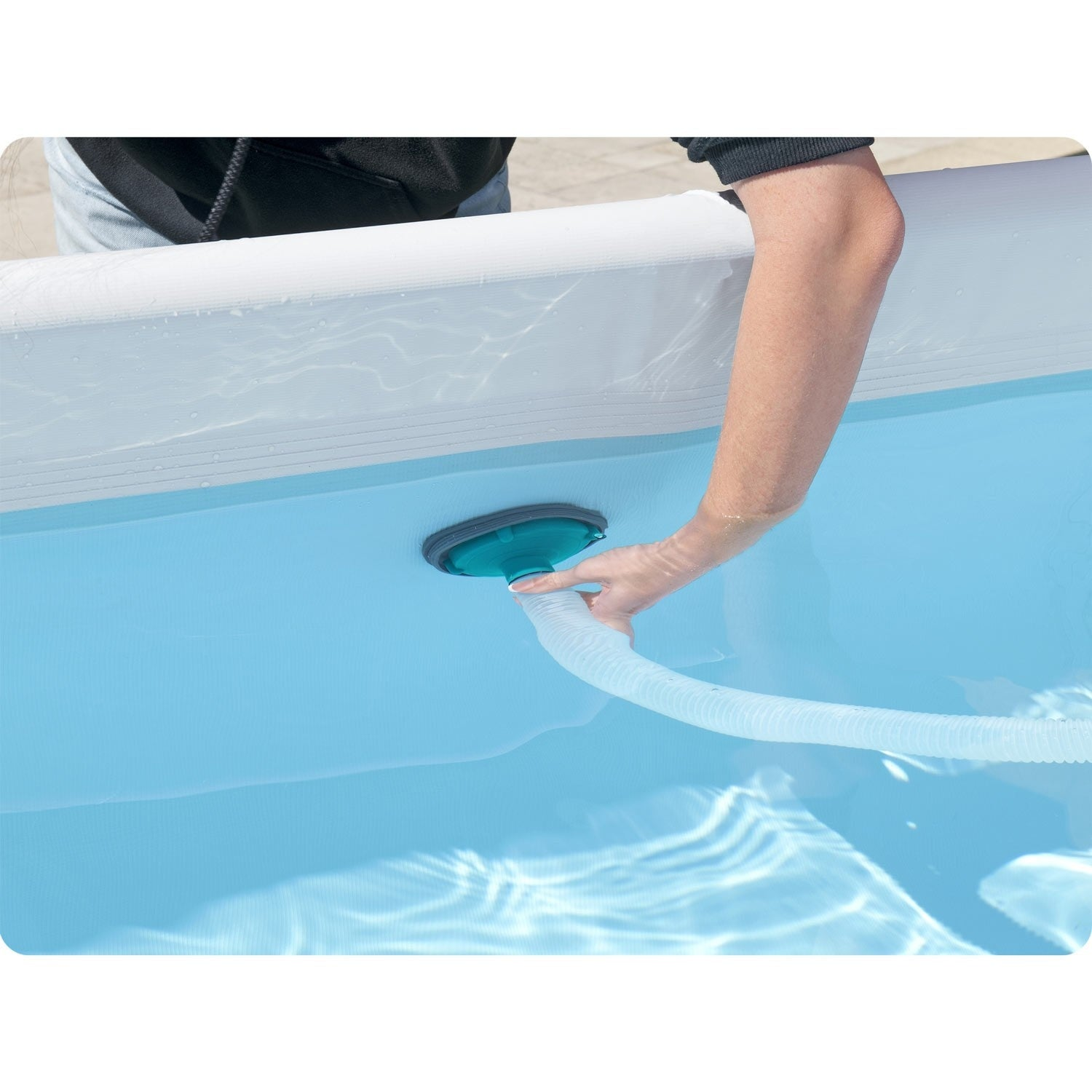 Una mano inserta una manguera en una piscina azul. El borde de la piscina es blanco. La persona lleva una camisa negra.