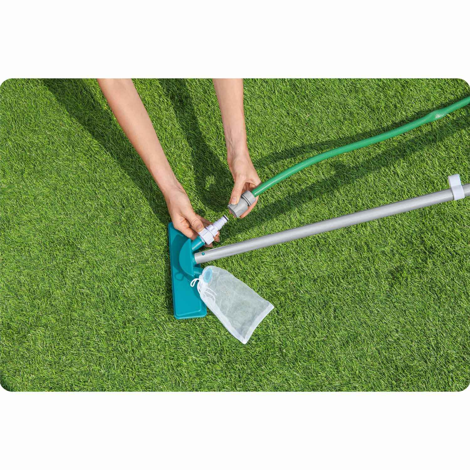 Manos conectan una manguera a un limpiador de piscina sobre césped verde. Hay una bolsa de malla blanca. La escena es al aire libre.