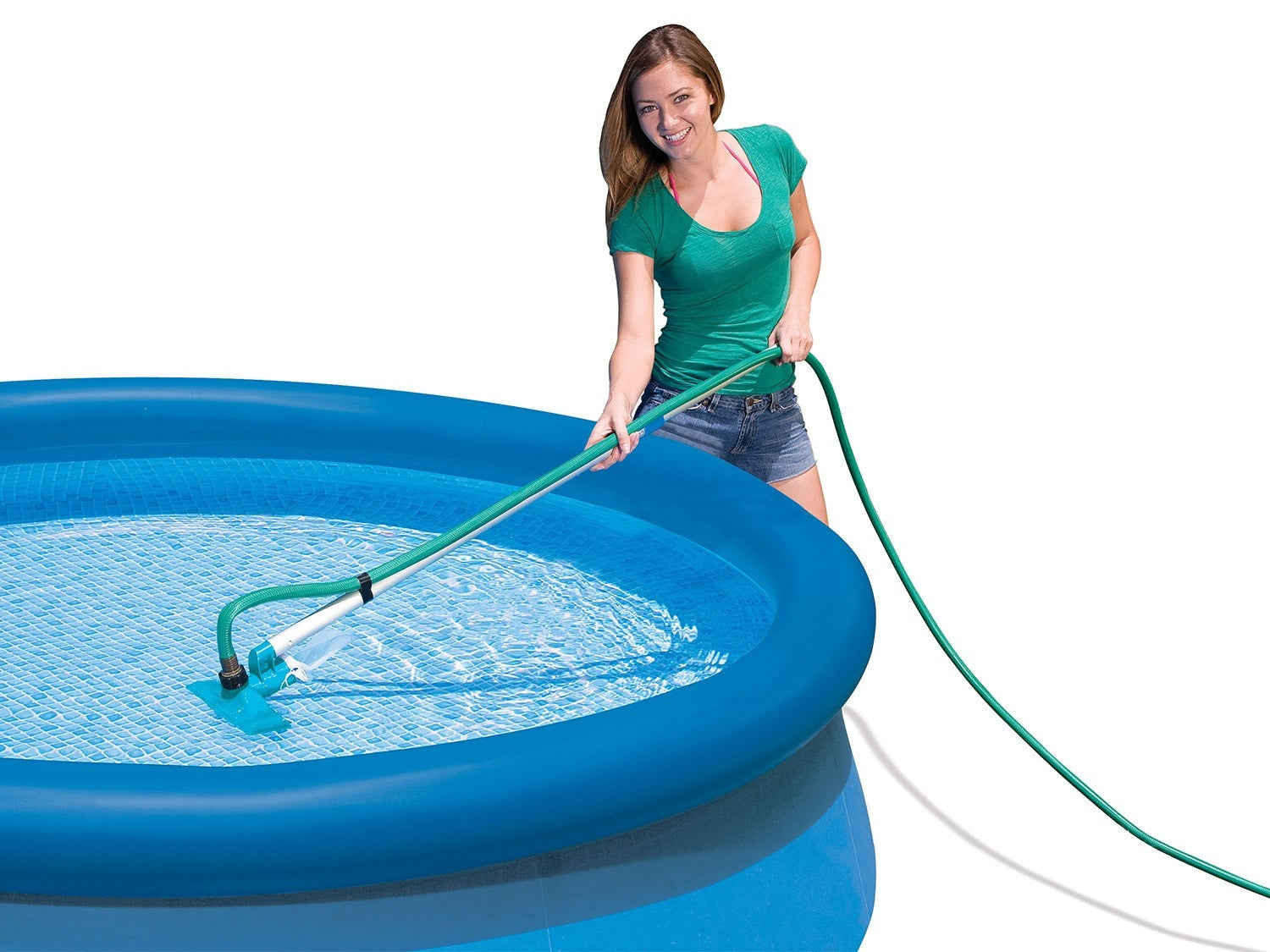 Una mujer usa una aspiradora de piscina en una piscina inflable azul. Lleva una camisa verde y pantalones cortos de mezclilla. El agua es clara.