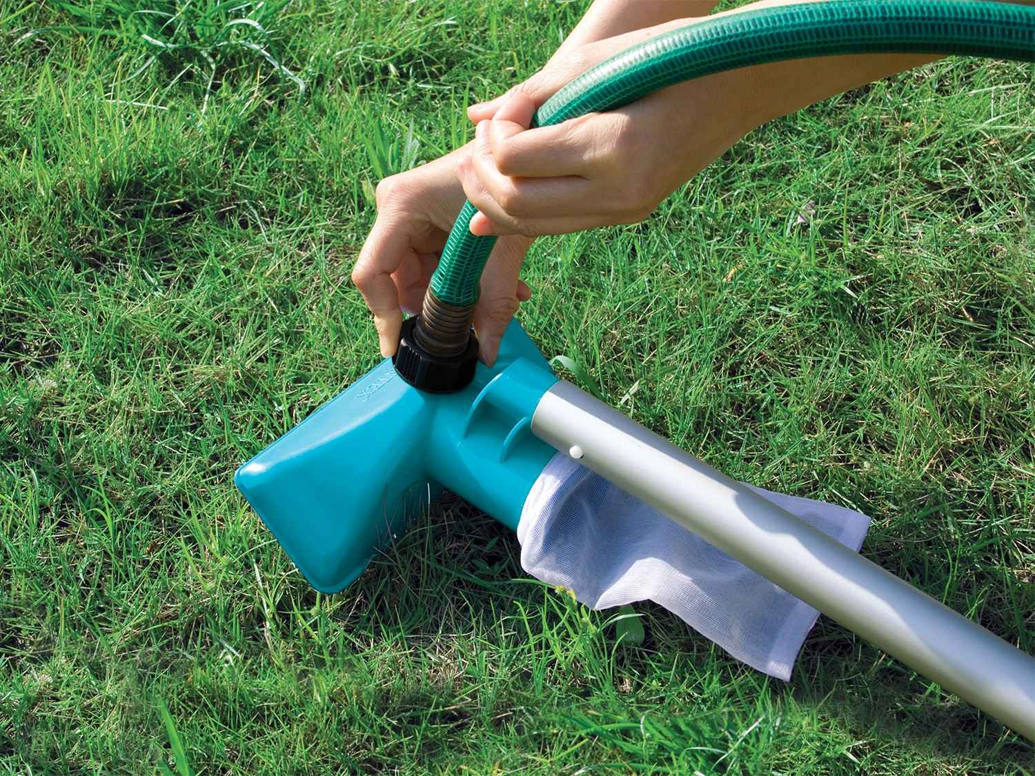 Manos conectando una manguera verde a una herramienta de jardín turquesa. La herramienta está sobre hierba verde, con una bolsa de malla blanca adjunta.
