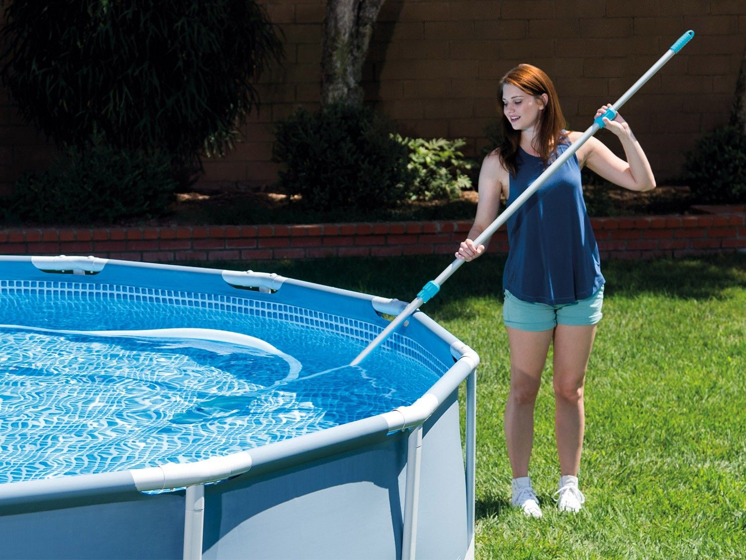 Una mujer usa un limpiador de piscina en una piscina azul elevada. Viste top y pantalones cortos azules, de pie sobre césped verde con árboles.