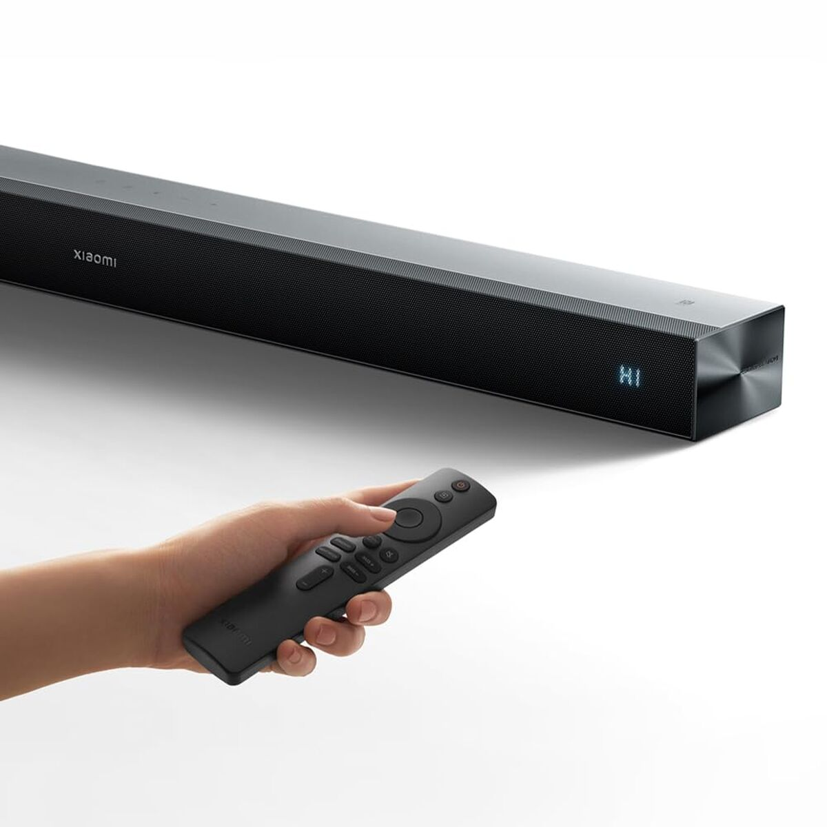 Eine Hand hält eine schwarze Fernbedienung, die auf eine schwarze Soundbar zeigt, die "HI" anzeigt. Auf der Soundbar steht "xiaomi".